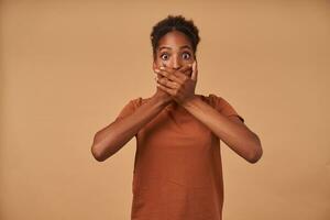 Shocked young attractive dark skinned brunette female covering her mouth with raised hands and rounding amazedly her brown eyes while standing over beige background photo