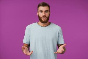 estudio Disparo de joven morena barbado masculino con corto Corte de pelo mirando a cámara perplejo y redondeo su ojos, posando terminado púrpura antecedentes con elevado palmas foto