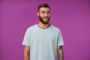 Beautiful astonished young tattooed male with beard looking aside with surprised face and showing his perfect white teeth, wearing casual clothes while standing over purple background photo