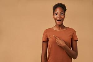 Bemused young pretty dark skinned curly woman looking surprisedly at camera with wide eyes and mouth opened while pointing aside with forefinger, isolated over beige background photo