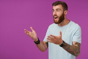 Portrait of young agitated brunette male with beard watching football on tv and being excited about game, looking aside with raised hands while posing over purple background photo