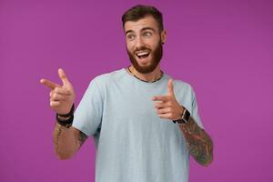 Joyful young attractive bearded man with short haircut raising index fingers and showing aside happily, contracting forehead and smiling with wide mouth opened, isolated over purple background photo