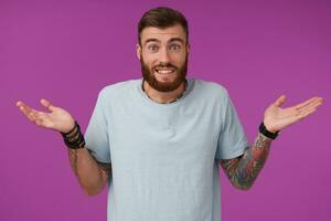 Portrait of young unshaved tattooed brunette man wearing blue t-shirt and trendy accessories contracting forehead and shrugging with raised palms, isolated over purple background photo