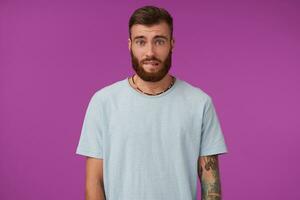 Beautiful young tattooed brunette male with beard looking guilty to camera and biting his underlip, keeping hands down while posing over purple background in casual clothes photo