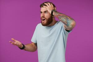 Studio shot of agitated tattooed brunette guy with beard clutching his head with raised hand and looking aside with confused face, isolated over purple background photo