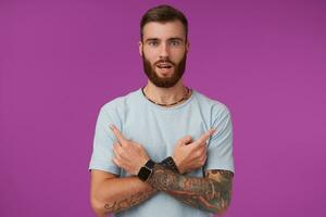 atractivo joven tatuado hombre con barba vistiendo azul camiseta y de moda accesorios, mirando a cámara y demostración en diferente direcciones con índice dedos, aislado terminado púrpura antecedentes foto