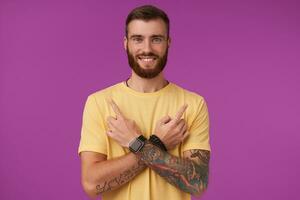 Cheerful pretty blue-eyed bearded male with tattooes wearing casual clothes and fitness watch posing over purple background, looking at camera with charming smile and keeping arms crossed photo