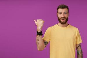 Attractive young tattooed brunette with trendy haircut looking at camera joyfully and smiling broadly, showing aside with raised thumb while standing over purple background photo