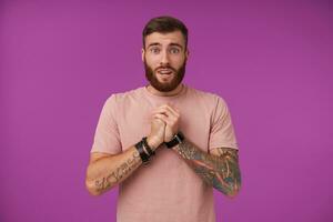 Portrait of pretty blue-eyed bearded brunette guy with tattooes raising folded hands and looking to camera pitifully, wearing beige t-shirt and trendy accessories while posing over purple background photo