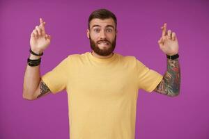 Hoping pretty bearded brunette male with tattooes making wish and crossing fingers, looking at camera and biting underlip, posing over purple background in yellow t-shirt photo