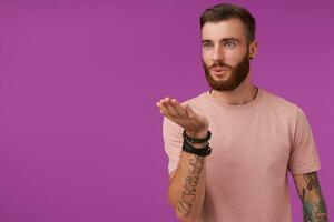 Good looking young bearded tattooed male with trendy haircut keeping palm raised and blowing air kiss aside, flirting with girl, posing over purple background in casual t-shirt photo