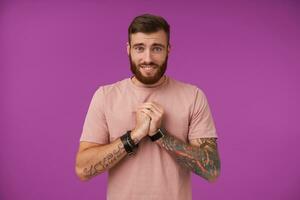 Pitiful attractive brunette man with beard and tattooes folding raised hands in begging gesture, wrinkling forehead and smiling broadly while looking to camera, isolated over purple background photo