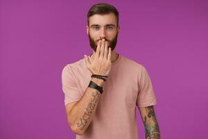 Charming young tattooed blue-eyed man with tattooes wearing beige t-shirt and trendy accessories while posing over purple background, keeping palm on his mouth and looking positively at camera photo