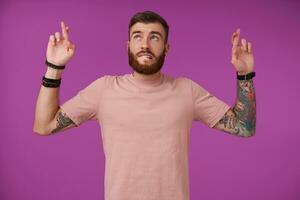 Portrait of handsome young bearded brunette male with tattooes and short haircut standing over purple background, looking upwards and biting underlip, crossing fingers and hoping for better photo