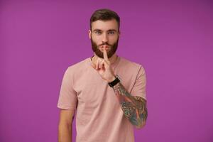 Good looking young bearded brunette man with tattooes keeping forefinger on lips in hush sign, asking to keep secret, wearing beige t-shirt and trendy accessories while posing over purple background photo