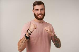 Portrait of handsome bearded tattooed guy with trendy haircut standing over white background in defensive position, looking at camera with charming wide smile photo