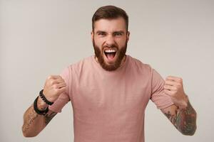 Studio shot of excited handsome bearded male with tattooes wearing beige t-shirt and trendy accessories, shouting loud and folding arms in fists while standing over white background photo