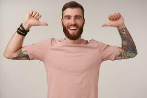 Happy attractive young bearded male with tattooes and short haircut wearing casual clothes while posing over white background, looking joyfully to camera and pointing to himself with raised thumbs photo