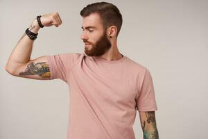 Studio shot of attractive bearded tattooed male having trendy haircut demonstrating his strong biceps, looking proudly on his raised hand, standing over white background photo