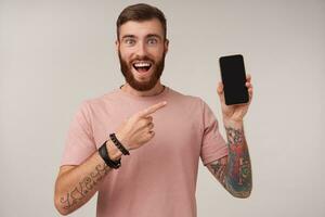 Shot of joyful young bearded brunette man with tattooes looking at camera with wide eyes and mouth opened and showing with forefinger on his smartphone in raised hand, isolated over white background photo