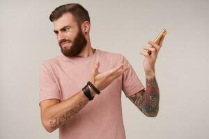 Disgruntled bearded tattooed man with short haircut keeping smartphone away from his ear, having unpleasant talk on phone, frowning his face and folding lips over white background photo