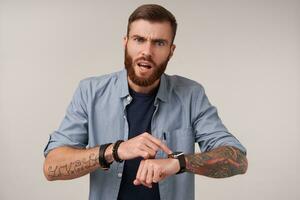 Grumpy blue-eyed brunette tattooed man with beard frowning his eyebrows while posing over white background, pointing to his watch and looking angrily to camera photo