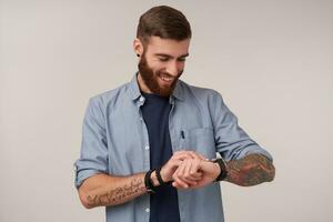 Studio shot of pleasant looking positive bearded man with short haircut standing over white background and setting time on his hand watch, being in good mood photo