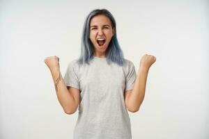 Overjoyed young attractive woman with short blue hair raising emotionally her hands and screaming happily with wide mouth opened, isolated over white background photo