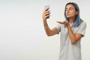Pleasant looking young tattooed lady with short blue hair keeping her eyes closed while blowing air kiss at camera of her smartphone, isolated over white background photo
