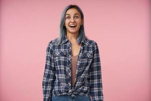 Bemused young lovely female with short blue hair looking excitedly at camera with wide eyes and mouth opened, dressed in checkered shirt while posing over pink background photo