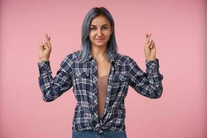 agradable mirando joven corto peludo mujer con natural maquillaje acuerdo su dedos cruzado y sonriente suavemente, vistiendo a cuadros camisa terminado rosado antecedentes foto