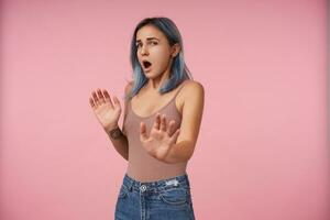 Studio photo of young pretty female with casual hairstyle raising hands in protective gesture while looking confusedly at camera, standing over pink background