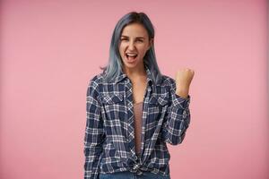 Joyful young attractive female with short blue hair raising joyfully her hand while looking exccitedly at camera, dressed in checkered shirt while posing over pink background photo