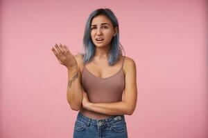 Portrait of bewildered young attractive short haired woman with tattoos raising confusedly palm while looking perplexedly at camera, isolated over pink background photo