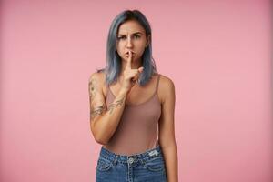 Indoor photo of young severe tattooed female keeping forefinger on her lips while looking seriously at camera, dressed in beige shirt while posing over pink background