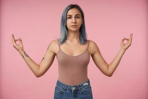 estudio Disparo de atractivo joven hembra con corto azul pelo plegable con elevado dedos namaste firmar mientras en pie terminado rosado antecedentes en beige camisa foto