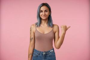 Positive young lovely tattooed lady with short blue hair smiling pleasantly while showing aside with raised hand, being isolated over pink background in casual wear photo