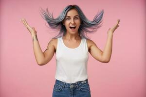 Agitated young pretty tattooed female with blue wild hair looking excitedly at camera with wide mouth opened and keeping her hands raised while posing over pink background photo