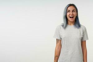 Joyful young lovely blue-haired woman with tattoos looking happily aside with opened mouth and keeping her hands down, isolated over white background photo