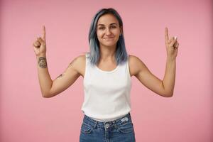 estudio Disparo de joven alegre tatuado hembra con corto Corte de pelo sonriente agradablemente mientras señalando hacia arriba con elevado índices, en pie terminado rosado antecedentes foto