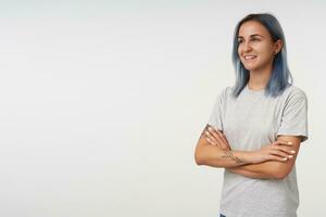 Side view of cheerful young pretty female with short blue hair crossing her hands on chest while looking positively ahead with pleasant smile, isolated over white background photo