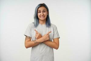 Confused young pretty tattooed blue haired woman keeping her arms crossed while showing in different sides and grimacing her face, posing over white background photo