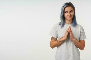 atractivo joven azul peludo mujer tatuajes plegable elevado manos y mirando astutamente aparte, vestido en gris básico camiseta mientras posando terminado blanco antecedentes foto