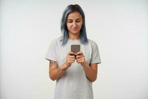 Positive young pretty blue-haired female with tattoos keeping mobile phone in raised hands while checking her messages, isolated over white background photo