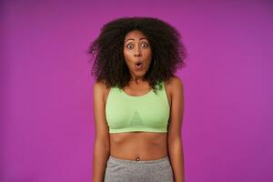 Shocked young dark skinned lady with curly hair posing purple background, keeping hands along the body, wearing light green top, looking at camera with round eyes and raising eyebrows photo