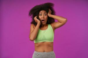 Studio shot of scared young dark skinned woman with curly hair keeping hands on her head and looking at camera with wide mouth opened, isolated over purple background photo