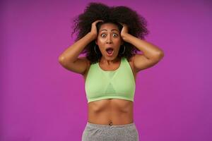 Portrait of astonished young dark skinned woman with curly hair standing over purple background, clutching her head and looking at camera with wide mouth opened, wearing casual clothes photo