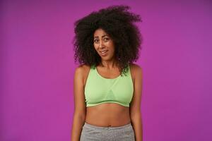 Indoor photo of confused young curly woman with dark skin wearing casual hairstyle, standing over purple background in sport clothes, looking at camera with puzzled face and wrinkling forehead