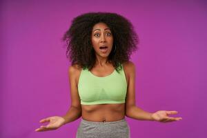 Indoor photo of young slim dark skinned female with curly hair wearing sporty clothes, raising palms up and looking at camera with amazed face, isolated over posing purple background