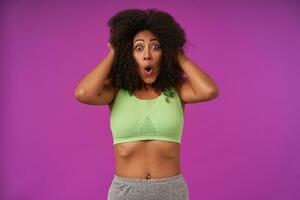 Amazed young curly dark skinned female wearing light green top, holding head with hands and looking at camera with round eyes and opened mouth, isolated over purple background photo